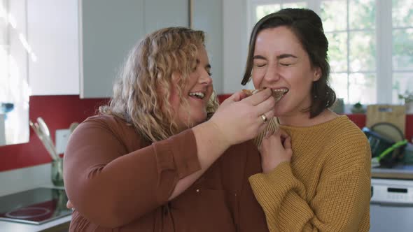 Happy caucasian lesbian couple eating bread and laughing in kitchen