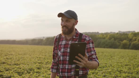 Smiled Bearded Man Talks on Tablet on Green Field