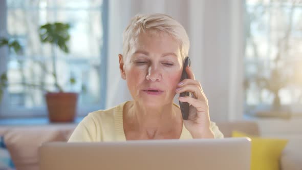 Elderly Woman Talking on Cellphone While Working on Laptop at Home