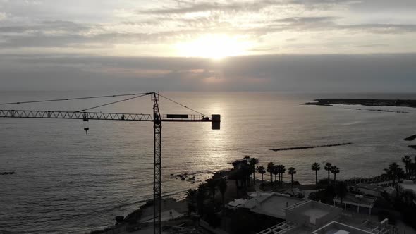 Aerial pan across a silhouette of a construction crane on the coast of Cyprus at sunset from right t