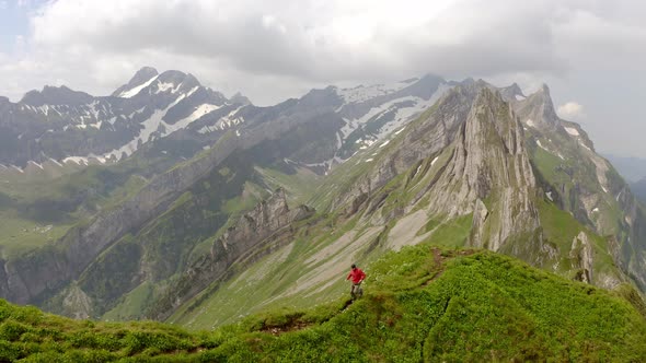 A man in a red jacket is running towards the camera on top of a mountain. There is a massive mountai