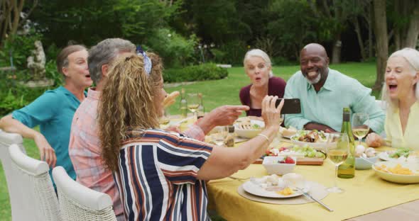 Animation of diverse happy senior female and male friends eating lunch in garden, using smartphone