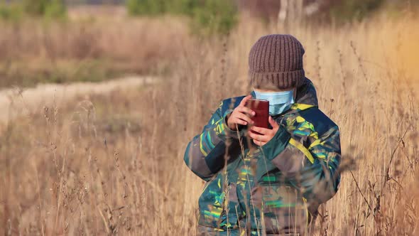 A Boy in a Mask on the Lawn Looks Attentively Into a Smartphone