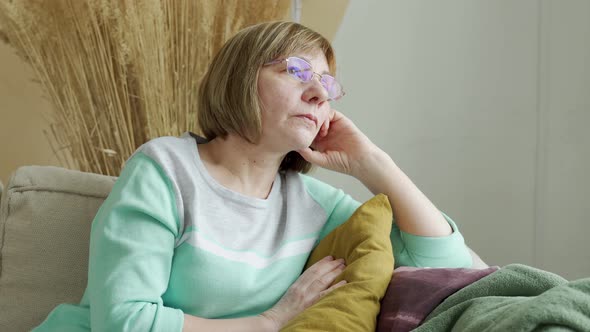 Thoughtful Elderly Woman Sits on the Sofa and Looks Out the Window