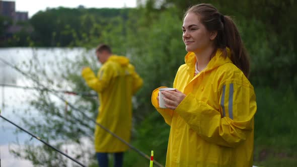 Beautiful Caucasian Young Woman Smiling Smelling Coffee in Cup As Blurred Man Adjusting Fishing Rod