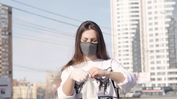 Young Woman on a City Background in a Medical Mask Puts on Sterile Gloves