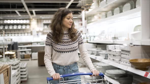 Young Woman Rolls Shopping Cart Along Shelves with Goods in Supermarket in Kitchen Department