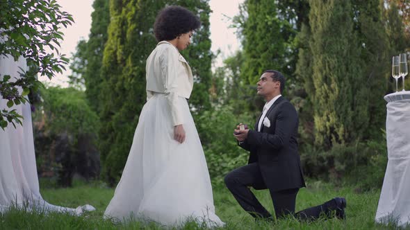 Happy Smiling African American Groom Standing on One Knee Proposing to Bride in Summer Garden at