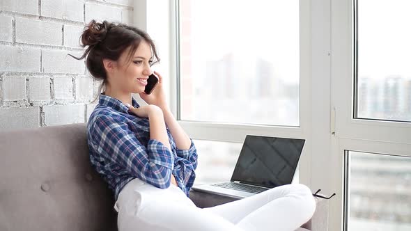 Attractive Woman Sitting on Couch Talking on Phone with Laptop at Home in Living Room