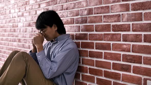 Christian Asian man is sitting against a wall, feeling sad, holding a cross and praying to God.