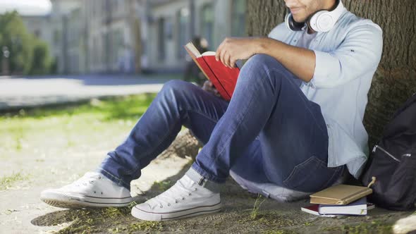 Multiracial Male Sitting Under Tree and Reading, Smiling and Closing Book, Happy