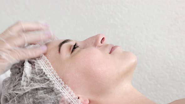 A female cosmetologist applies cream to the client's face with massaging movements.