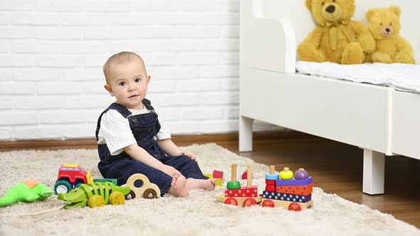 a Baby in a Denim Jumpsuit Sitting Barefoot on the Carpet Smiles and Plays with Toys