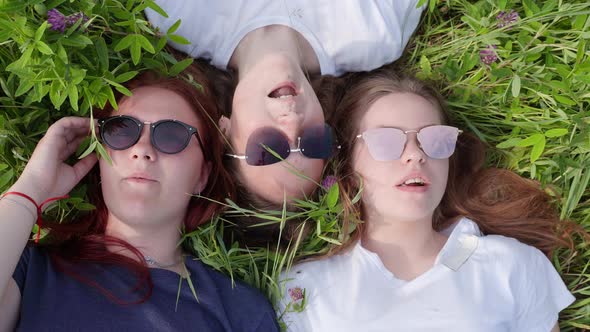 Three Friendly Girlfriends Are Lying on the Grass with Sunglasses and Chewing Gum.