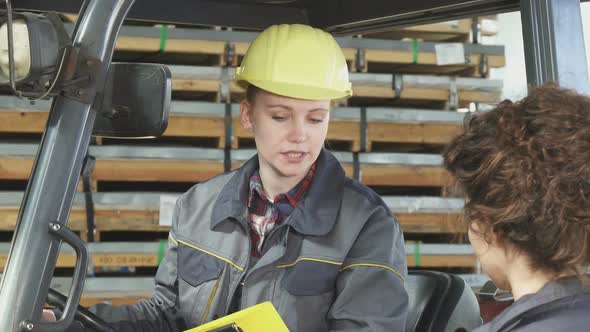 Young Female Fatory Worker Operating Forklift at the Storage Talking To Her Colleague