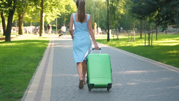 Closeup of Young Woman Walking on City Sidewalk with Green Suitcase on Summer Day