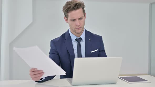 Young Businessman Reading Documents and Working on Laptop
