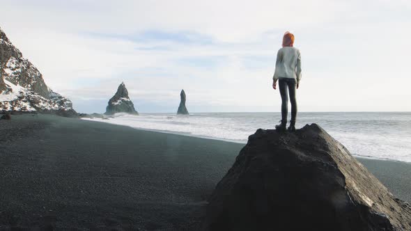 Beautiful Redhaired Woman Traveler Standing on a Huge Stone on Volcanic Black Sand Beach in Iceland