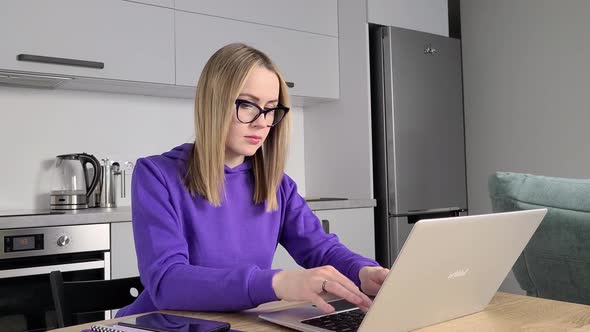 Pensive Young Adult Woman in Glasses Work Study at Kitchen Using Laptop