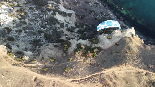 Aerial Drone View of a Man Flying a White and Blue Paraglider Over a Hill and Trees to the Sea Waves