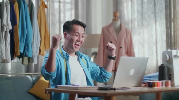 Happy Asian Male Designer Working On A Laptop And Celebrating And Dancing While Designing Clothes