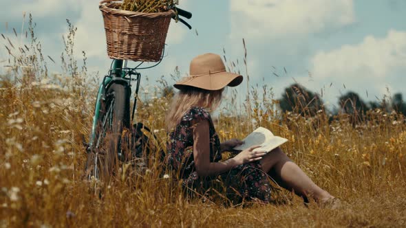 Woman Sitting On Flower Meadow And Reading Book Diary. Tourist Girl Relaxing On Wildflower Field.