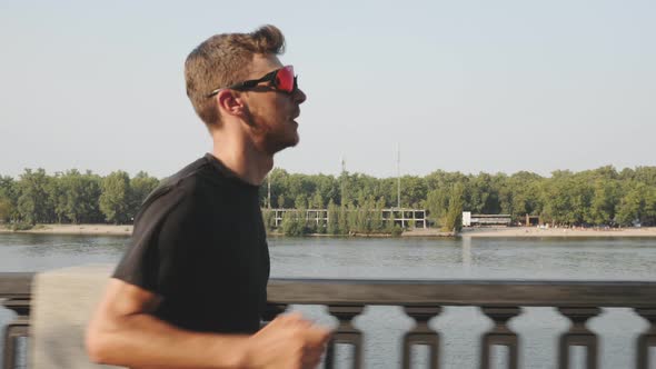 Strong focused young man in black sportswear runs along city river at sunset training for marathon