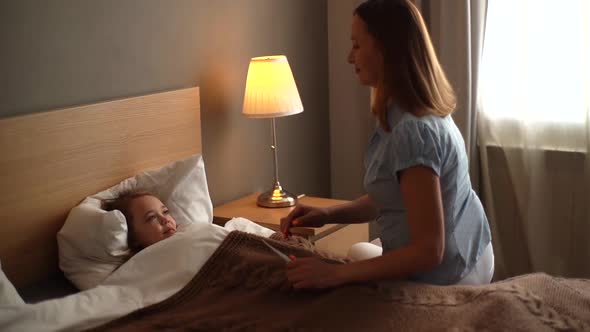 Loving Mother Touches Forehead of Cheerful Recovering Daughter Lying in Bed and Measures Temperature