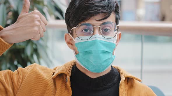 Closeup Young Indian Guy in Medical Mask Posing Indoors Looking at Camera Showing Thumbs Up Sign