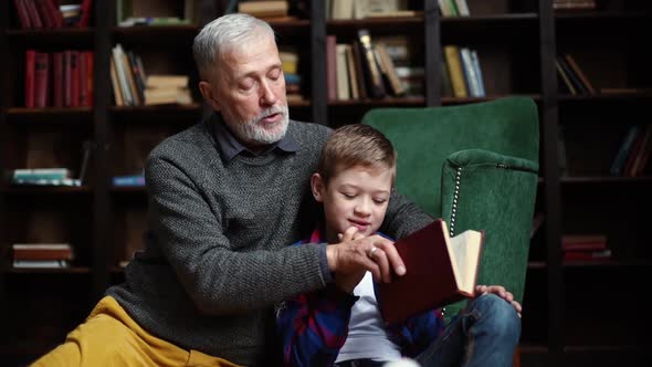 Portrait of Little Boy Sitting on Knees of His Grandfather and Reading an Interesting Book Together