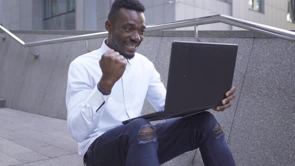 Portrait of Handsome African American Man in White Shirt Typing on Laptop Sitting on The City Street