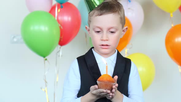 Boy in Party Cone Blows Candle on Cupcake Against Balloons