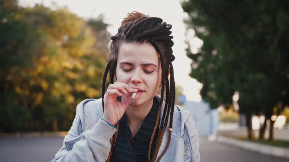 Portrait of Young Pretty Woman with Dreadlocks Smoking Marijuana Joint in Park Slow Motion