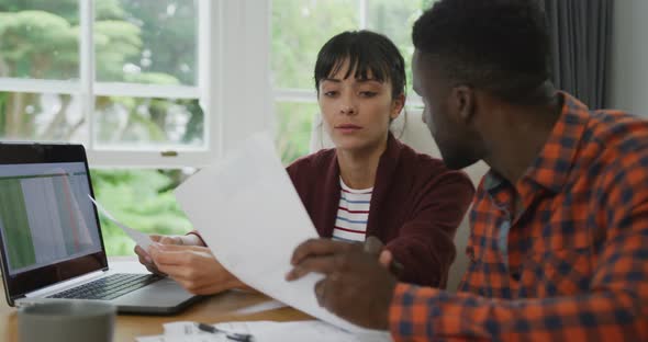 Diverse couple sitting at table talking and working with laptop