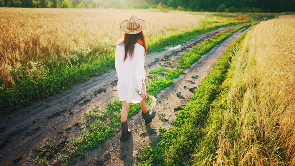 Rear View of Beautiful Girl in White Dress and Straw Hat Strolling Through Field Holding Bouquet of