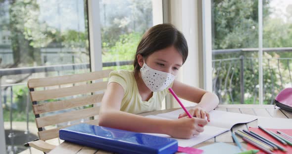 Caucasian girl spending time at home wearing face mask, sitting at table doing her homework, in slow