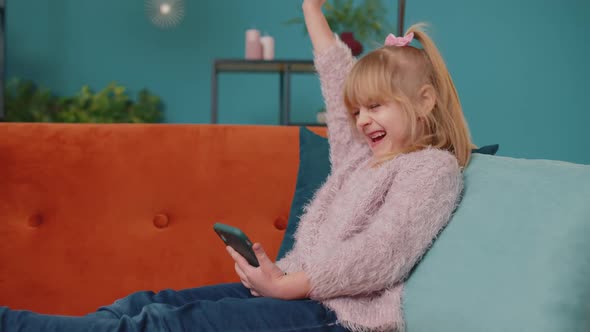 Happy Little School Kid Girl Use Mobile Cell Phone Doing Winner Gesture Celebrate Alone at Home Sofa