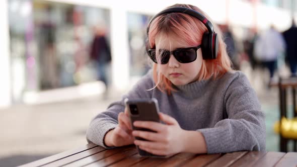 Cute Child Girl in Headphones and Moms Sunglasses is Playing Game in Mobile Phone in Cafe