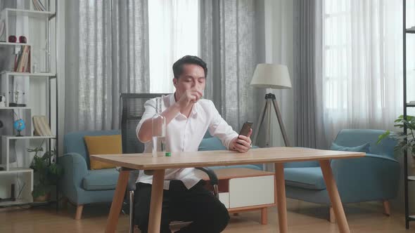 Drunk, Depressed Asian Man Drinking Vodka From Glass During Using Smartphone At Home