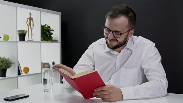 Smart Smiling Bearded Man in White Shirt, Glasses Reading Printed Book at Home