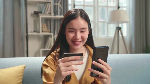 Asian Woman Sitting On Couch In Living Room Holding Credit Card And Using Smartphone Shopping Online