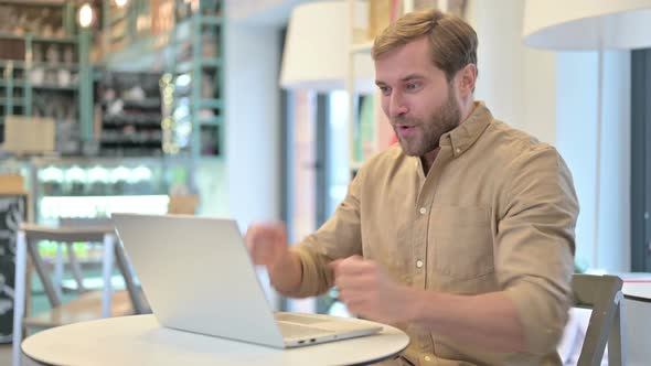 Young Man Celebrating Success on Laptop in Cafe