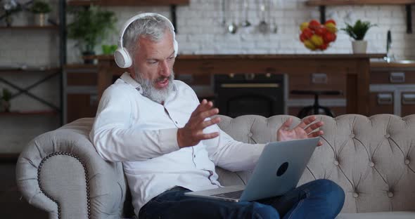 Side View Bearded Mature Business Man in Headphones Talking Video Call at Home Sitting on the Sofa