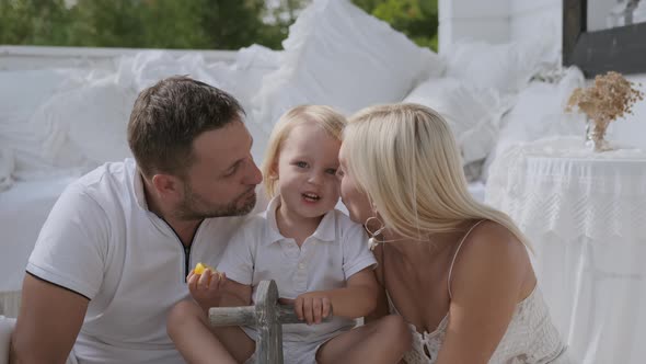 Mom and Dad Kiss Their Little Son From the Cheek on Both Sides Sitting on the Porch of the House