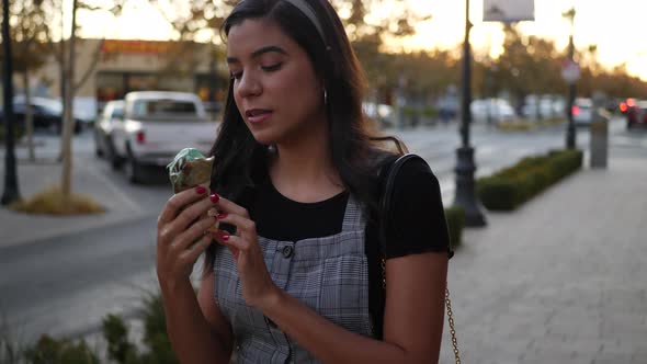 A cute young hispanic woman eating a messy, dripping ice cream cone walking on a city street.
