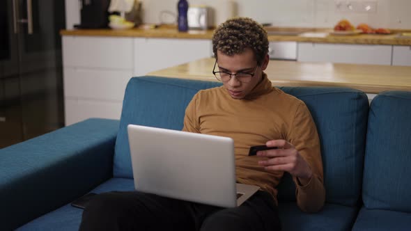 Young Man Buying Online Uses a Laptop and a Credit Card on Sofa Home