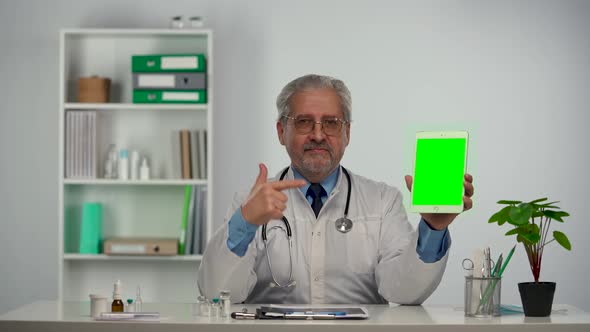 Aged Male Doctor Holds Tablet with Green Screen Chroma Key Points at It with Finger and Makes Thumb