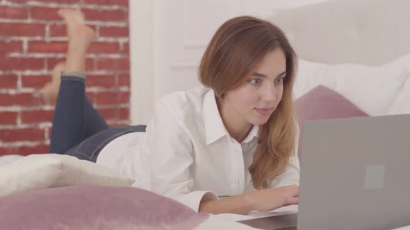 Smiling Caucasian Girl Laying in Bed with Laptop and Typing