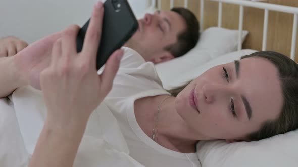 Beautiful Woman Using Smartphone Next To Sleeping Partner in Bed 