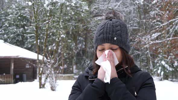 Young asian woman blows her nose in cold winter forrest in Switzerland.
